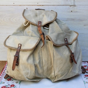 Può includere: Uno zaino in tela beige caratterizzato da un ampio scomparto principale con chiusura a patta, due tasche frontali con patta e cinturini in pelle marrone con fibbie in metallo. La borsa presenta cuciture e rivetti in metallo a vista.