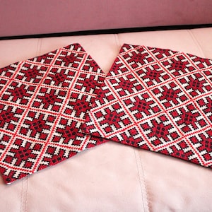 May include: Two square cushions with a geometric pattern in red, black, and white. The cushions are placed side by side, showcasing the intricate embroidered design. The cushions are likely used for home decor.