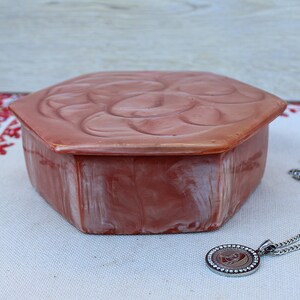 May include: A pink hexagonal trinket box with a floral design on the lid. The box is sitting on a white tablecloth with red embroidery. A silver chain necklace with a round pendant is next to the box.