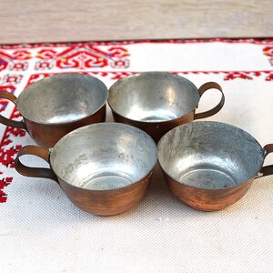 May include: Four vintage copper and metal cups with handles. The cups have a copper exterior and a silver-coloured interior. They are arranged on a white tablecloth with a red and white patterned border. The cups are likely for serving beverages.