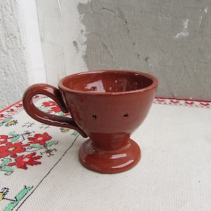 Könnte beinhalten: Eine Terrakotta-Tasse mit Henkel und Löchern im Boden. Die Tasse steht auf einer weißen Tischdecke mit rotem und grünem Blumenmuster.