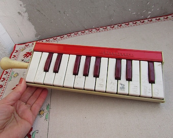 May include: A vintage red and white toy piano with a cream-colored mouthpiece. The keys are white with dark red accents. The word "Simba" is printed in white on the red top. Some keys have handwritten markings.