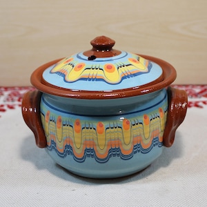 Może przedstawiać: Ceramiczny garnek z pokrywką, z jasnoniebieską podstawą i pokrywką z brązową obwódką i uchwytami. Garnek zdobiony jest kolorowym, powtarzającym się wzorem w kolorze żółtym, pomarańczowym i czarnym. Pokrywka ma mały brązowy uchwyt.