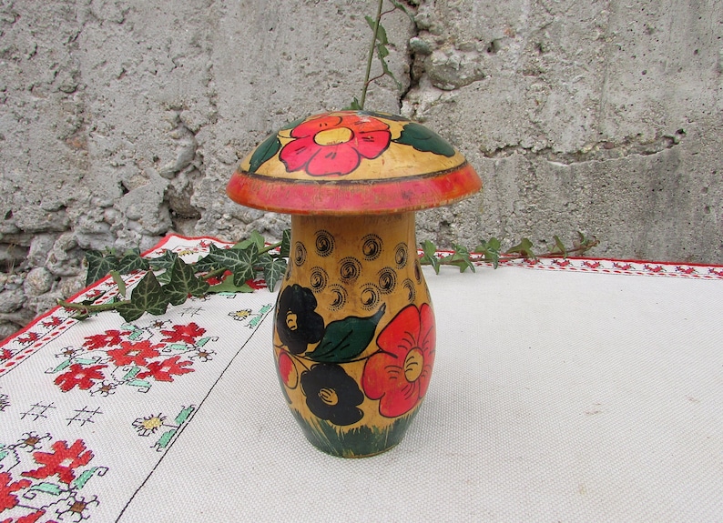 May include: A wooden mushroom-shaped container with a red, yellow, and black floral design. The container is painted with a yellow background and has a red rim.