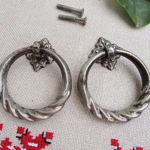 Op de afbeelding: Twee zilveren ringdeurgrepen met een gedraaid touwontwerp en sierlijke bloemmotieven.