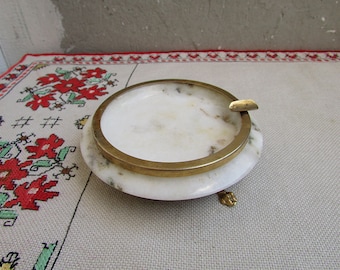 May include: A white alabaster ashtray with a gold rim and three gold feet. The ashtray has a slight marbling effect.