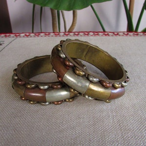 Pode incluir: Dois braceletes de latão com um design decorativo de bandas alternadas de cobre, prata e latão. Os braceletes estão empilhados um em cima do outro.