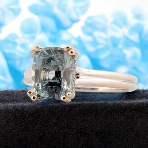 K&ouml;nnte beinhalten: Ein silberner Ring mit einem gr&uuml;nen Kissen-Schliff-Edelstein, der in einer goldenen Fassung gefasst ist. Der Ring liegt auf einem schwarzen Samt-Untergrund.