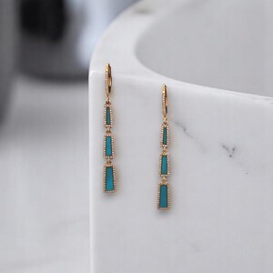 May include: Dangling earrings with a gold-colored metal setting. Each earring features three rectangular turquoise-colored stones. The earrings have a lever back closure and are displayed against a white background.