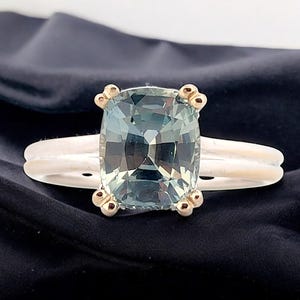 K&ouml;nnte beinhalten: Ein silberner Ring mit einem gr&uuml;nen Edelstein im Kissen-Schliff, der in einer goldenen Fassung sitzt. Der Ring hat eine doppelte Band und goldene Akzente.