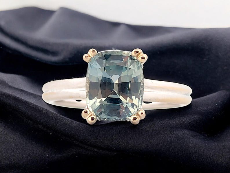 K&ouml;nnte beinhalten: Ein silberner Ring mit einem gr&uuml;nen Edelstein im Kissen-Schliff, der in einer goldenen Fassung sitzt. Der Ring hat eine doppelte Band und goldene Akzente.