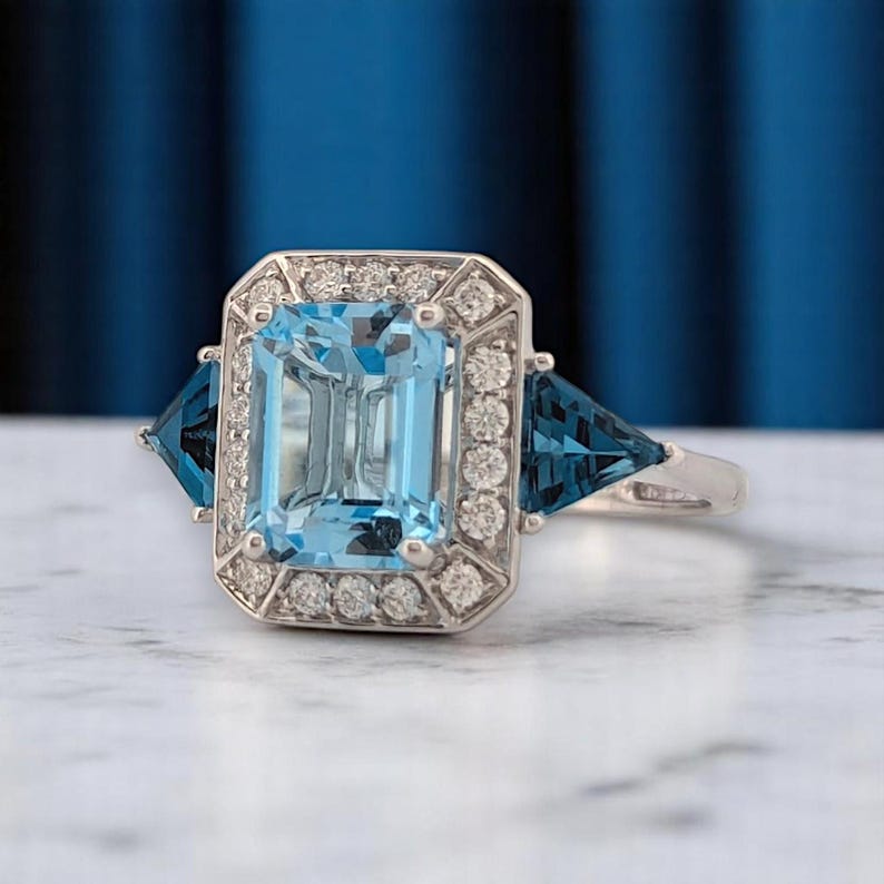 Puede incluir: Un anillo de plata con una gran gema rectangular azul claro, rodeada de piedras blancas m&aacute;s peque&ntilde;as. Dos gemas triangulares azul oscuro flanquean la piedra central. El anillo se encuentra sobre un fondo azul borroso.