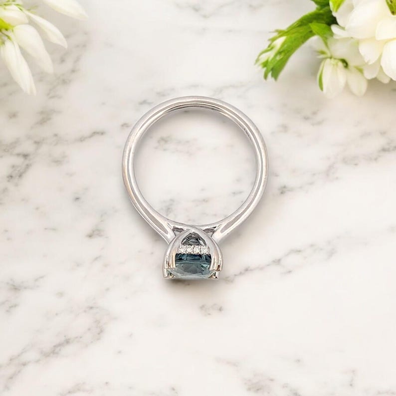 K&ouml;nnte beinhalten: Ein silberner Ring mit einem blauen Edelstein, der in einer Krappenfassung sitzt. Der Edelstein ist von einem Heiligenschein aus kleineren Diamanten umgeben.