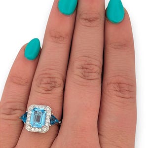 Puede incluir: Un anillo de plata con una gran piedra preciosa rectangular azul claro, rodeada de peque&ntilde;as piedras transparentes. Dos piedras preciosas azules triangulares flanquean la piedra central. El anillo est&aacute; en una mano con esmalte de u&ntilde;as turquesa.