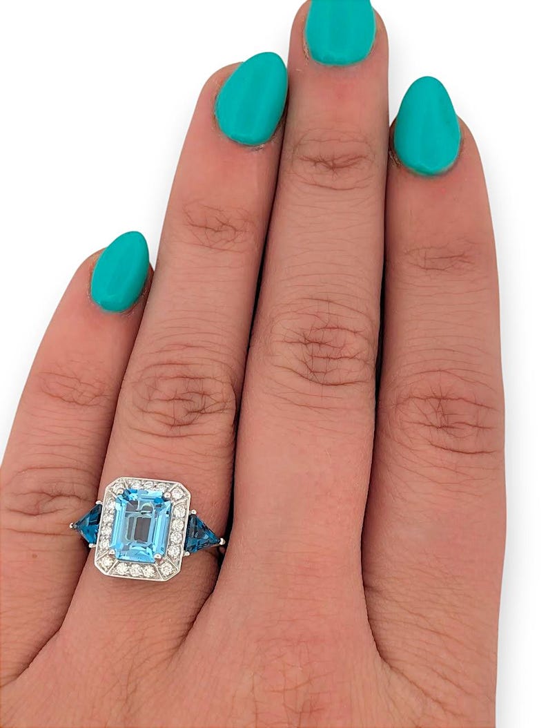 Puede incluir: Un anillo de plata con una gran piedra preciosa rectangular azul claro, rodeada de peque&ntilde;as piedras transparentes. Dos piedras preciosas azules triangulares flanquean la piedra central. El anillo est&aacute; en una mano con esmalte de u&ntilde;as turquesa.