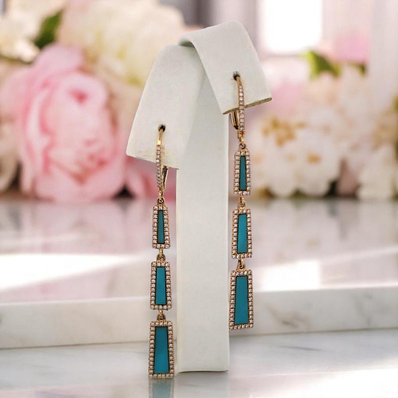 May include: Gold-toned dangle earrings with rectangular turquoise-colored insets, framed by a border of small, sparkling stones. The earrings are displayed on a white jewelry stand, with a soft floral background.