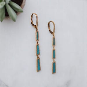 May include: A pair of gold-toned drop earrings. Each earring features three turquoise-colored rectangular stones set in a gold frame, with a small, diamond-like accent at the top. The earrings are displayed on a white surface.