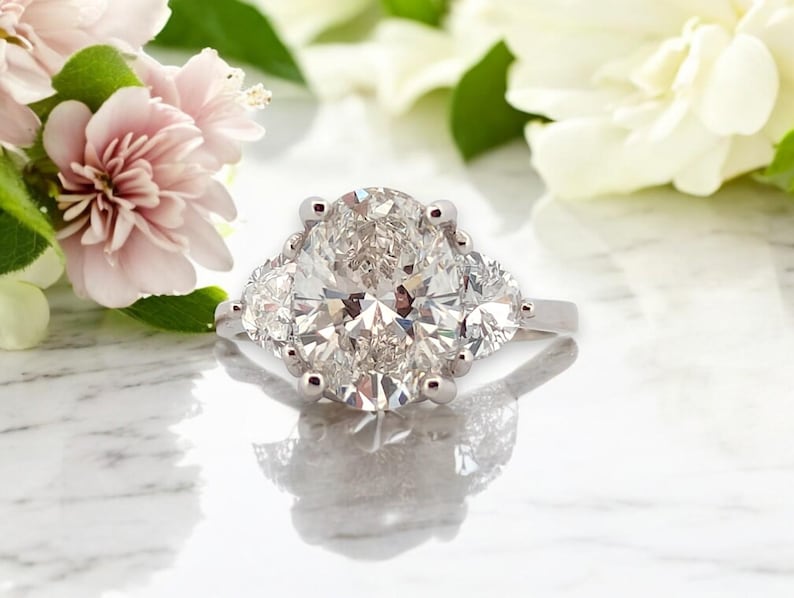 May include: A silver engagement ring with a large oval-shaped diamond surrounded by two smaller diamonds on either side.