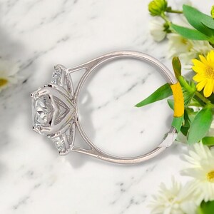 May include: A silver ring with a large, square-shaped diamond in the center and two smaller diamonds on either side. The ring is set on a white marble surface.