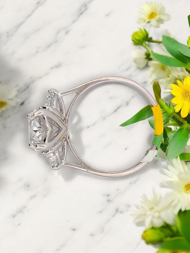 May include: A silver ring with a large, square-shaped diamond in the center and two smaller diamonds on either side. The ring is set on a white marble surface.