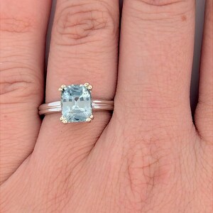 K&ouml;nnte beinhalten: Ein silberner Ring mit einem quadratischen, hellblauen Edelstein, der in einer goldenen Fassung sitzt. Der Ring hat ein d&uuml;nnes Band mit einer dezenten Textur.