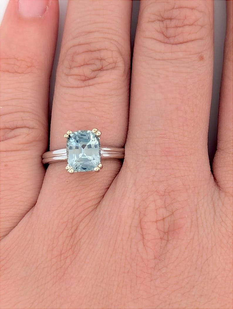 K&ouml;nnte beinhalten: Ein silberner Ring mit einem quadratischen, hellblauen Edelstein, der in einer goldenen Fassung sitzt. Der Ring hat ein d&uuml;nnes Band mit einer dezenten Textur.