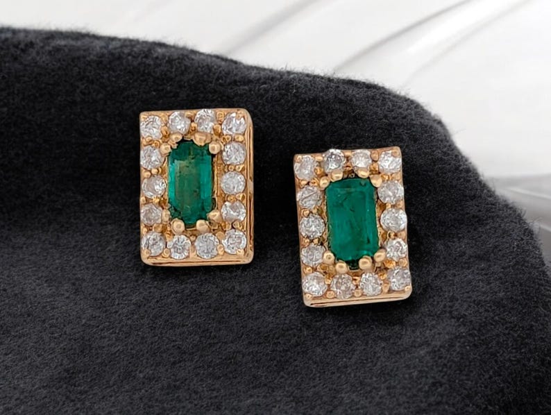 Op de afbeelding: Een paar gouden oorbellen met smaragdgroene edelstenen omringd door kleine heldere diamanten.