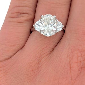 May include: A three-stone diamond engagement ring with a large oval-shaped center stone and two smaller pear-shaped stones on either side. The ring is set in platinum.