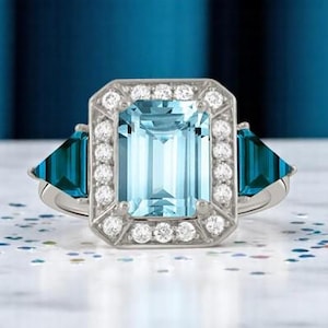 Puede incluir: Un anillo plateado con una gran gema rectangular azul claro, rodeada de piedras blancas m&aacute;s peque&ntilde;as. Dos gemas triangulares de color turquesa flanquean la piedra central. El anillo se encuentra sobre un fondo azul borroso.