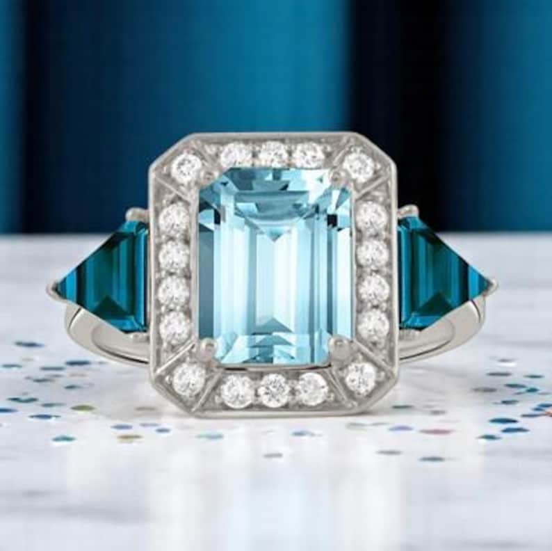 Puede incluir: Un anillo plateado con una gran gema rectangular azul claro, rodeada de piedras blancas m&aacute;s peque&ntilde;as. Dos gemas triangulares de color turquesa flanquean la piedra central. El anillo se encuentra sobre un fondo azul borroso.