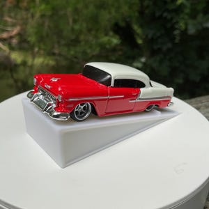 May include: A die-cast toy car featuring a two-tone red and white exterior paint scheme. The vehicle has a chrome-colored front grille, bumper, and side trim accents. It is displayed on a white angled stand, showcasing its classic body style.