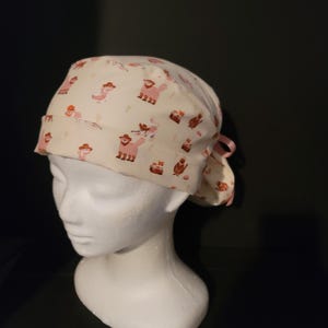 May include: A white scrub hat with a pink and brown pattern of small animals. The hat has a tie on the back.