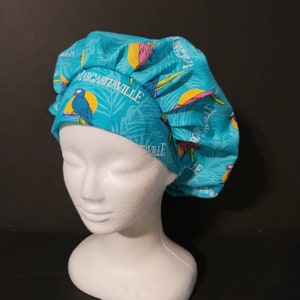 May include: A turquoise bouffant scrub cap with a tropical print featuring parrots, palm leaves, and the text "Margaritaville." The cap is displayed on a white mannequin head against a dark background.