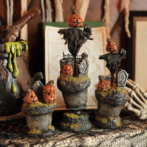 May include: Halloween-themed decorative figurines. The set includes three pumpkin-headed scarecrows atop mossy, textured bases. Each figure has a carved pumpkin face and is adorned with a black, tattered outfit. Tombstones and a cauldron are also visible.