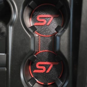 May include: A pair of circular black cup holder inserts with a textured surface and a red border. Each insert features a prominent red ST logo in the center. The inserts are positioned within a black plastic automotive center console cup holder.