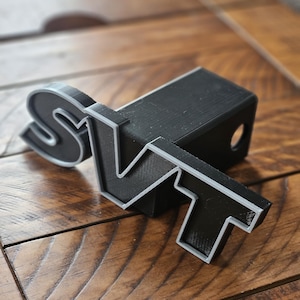 May include: Black plastic hitch cover with a silver SVT logo. The logo is raised and has a 3D effect.