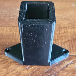 May include: A black 3D printed plastic stand with a square opening on top and a hexagonal base.