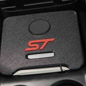 May include: A black textured console insert featuring a prominent red ST logo, a circular black accent, and a silver metallic rectangular detail. The insert is designed to fit into a vehicle center console area with integrated cup holders.