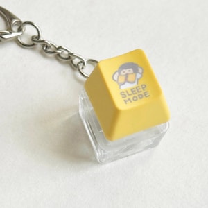 Puede incluir: Un llavero amarillo con forma de tecla de teclado con las palabras "SLEEP MODE" y un dibujo de un mono. El llavero tiene una cadena y un cierre plateados. La tecla está montada sobre una base cuadrada transparente.