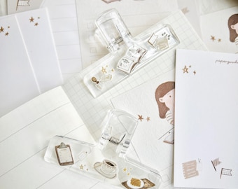 May include: A collection of stationery items, including notepads, paper clips, and decorative paper. The paper clips are clear with illustrations of a book, coffee cup, and food. The notepads and paper feature illustrations of a girl, stars, and other small graphics.
