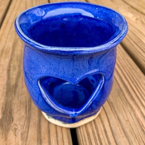 May include: A blue ceramic oil burner with a heart-shaped cutout. The burner is sitting on a wooden surface.