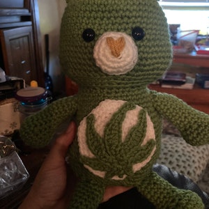 Könnte beinhalten: Grüner, gehäkelter Stoffbär mit einem weißen und grünen Marihuana-Blatt-Design auf der Brust. Der Bär hat schwarze Augen und eine weiße Nase mit einem gelben Herzen.