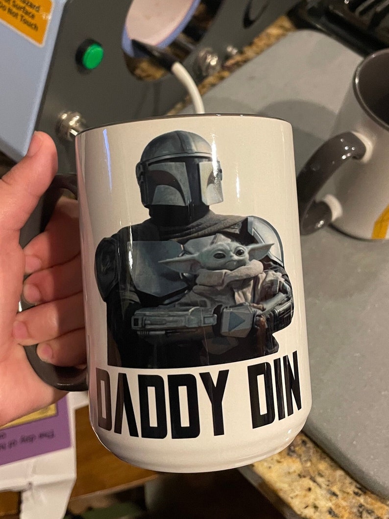 Puede incluir: Taza de cer&aacute;mica blanca con asa negra. La taza presenta un gr&aacute;fico de un personaje mandaloriano sosteniendo un peque&ntilde;o alien&iacute;gena verde. El texto "Daddy Din" est&aacute; impreso en negro debajo del gr&aacute;fico.