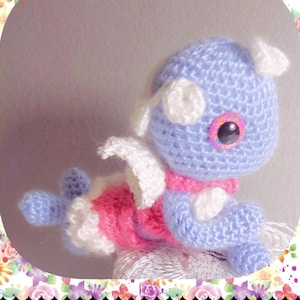 May include: A crocheted plush toy in the shape of a dragon. The dragon is light blue with pink and white accents. It has a large, white eye with a black pupil and a pink iris.