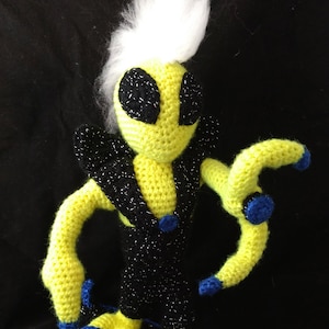 May include: A yellow crocheted alien with a white fuzzy mohawk, wearing a black and sparkly outfit, riding a black and blue surfboard.