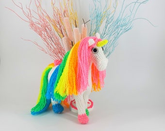 May include: A white crocheted unicorn with a rainbow mane and tail. The unicorn is standing on a white cake with pink and green frosting. The unicorn is surrounded by pink, yellow, and blue branches.