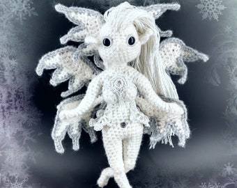 May include: A white crocheted fairy with gray wings and long white hair. The fairy has large black eyes and a white crocheted dress with a snowflake design. The fairy is standing on a dark background with a snowflake pattern.