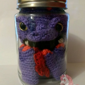 May include: A purple crocheted creature with red accents is inside a clear glass jar with a silver lid. The creature has large black eyes with gold accents. The jar is sitting on a white surface.