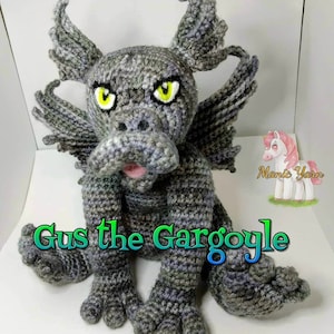 May include: A gray crocheted gargoyle with green eyes and a pink tongue. The gargoyle is sitting on a white surface and has the text "Gus the Gargoyle" written in green and blue letters.