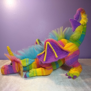 May include: A rainbow striped crocheted dragon with a purple and yellow wing, blue fur, and a yellow tail.